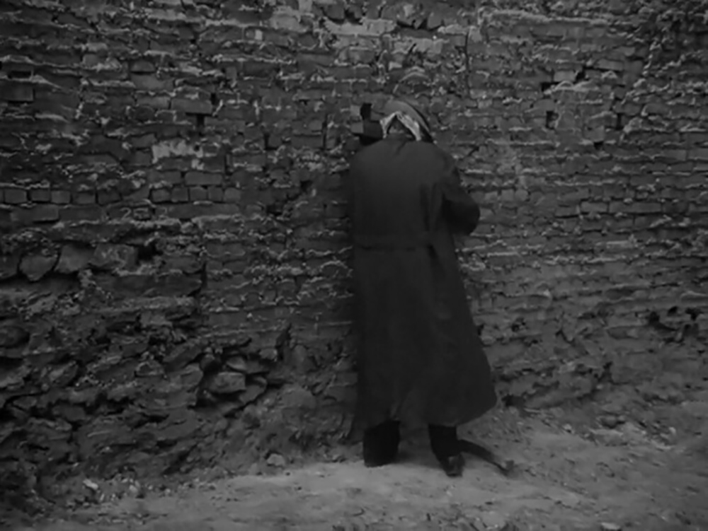 O standing against a stone wall while concealing themself from E in pursuit of an androgynous state.